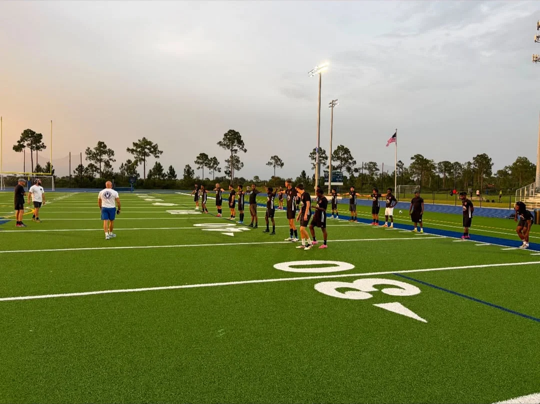 Q Fitness coaches running a warm-up with the SWFL SC Swamp Dragons squad on the field at dusk,
                    stadium lights on and the team lined up across the 30-yard line.